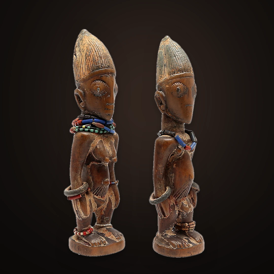 Dos figuras Ibeji de madera, con cofias elevadas y collares de cuentas, vistas en tres cuartos